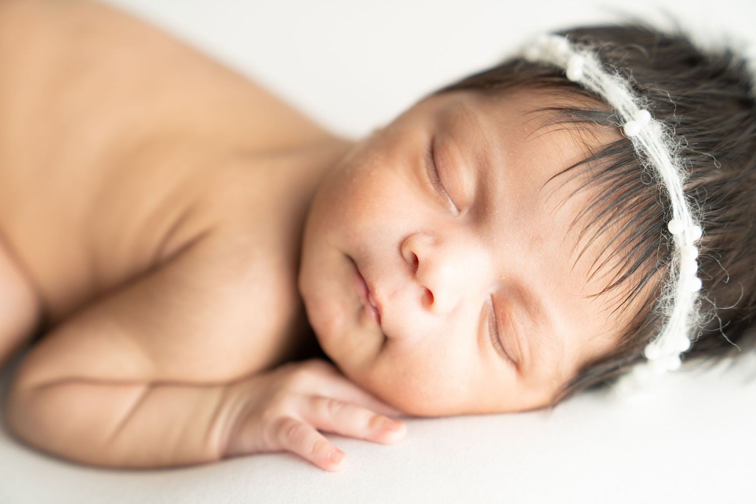 Peaceful newborn baby girl sleeping with a delicate white floral headband on a soft white background.