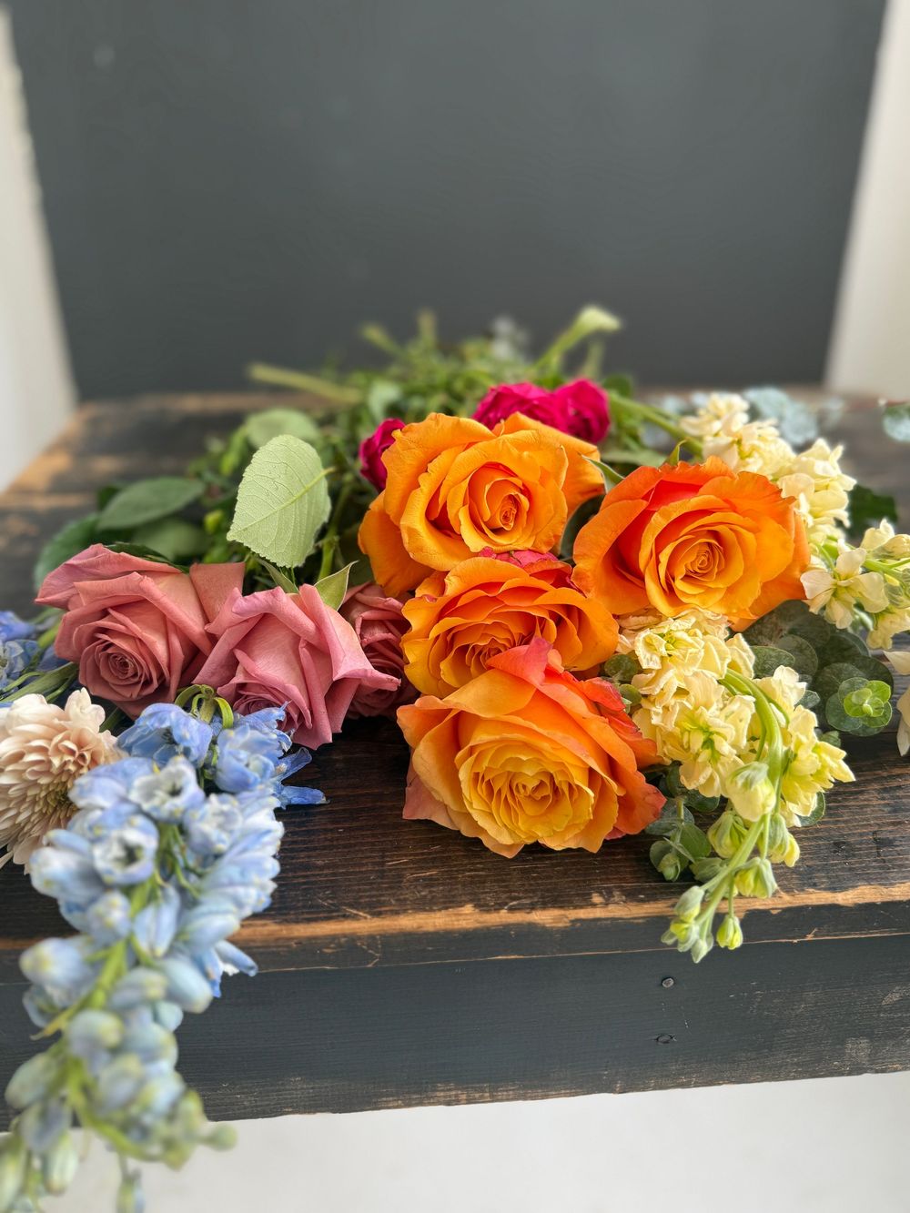 Colorful floral arrangement with orange roses, pink roses, blue delphinium, and yellow stock flowers on a rustic wooden box.