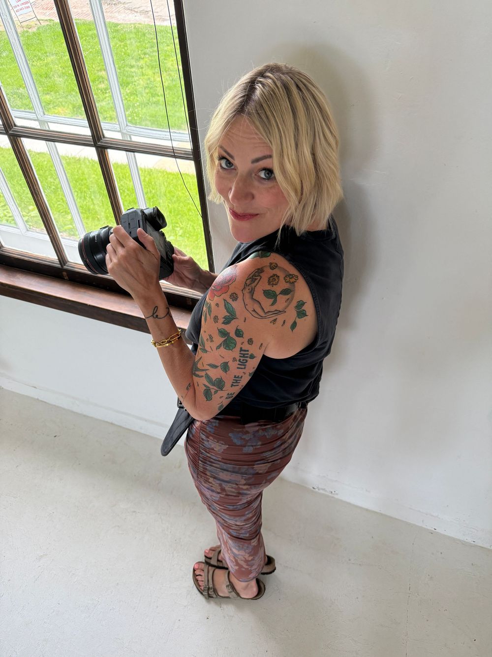Blonde woman with arm tattoo holding a camera near a window, wearing a black top and patterned leggings.