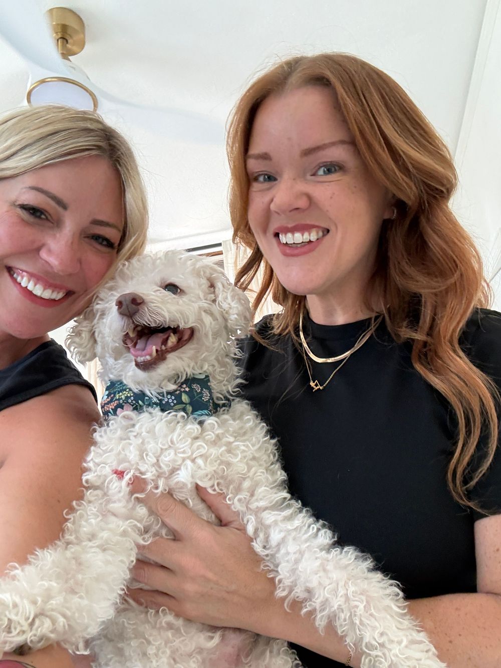 Two smiling women holding a fluffy white dog, posing together for a cheerful selfie indoors.