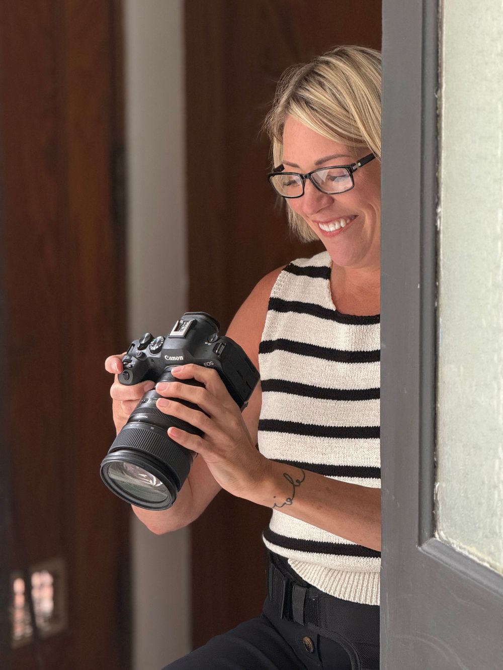 Smiling blonde woman with glasses holding a Canon DSLR camera, wearing a striped top, standing in a doorway.