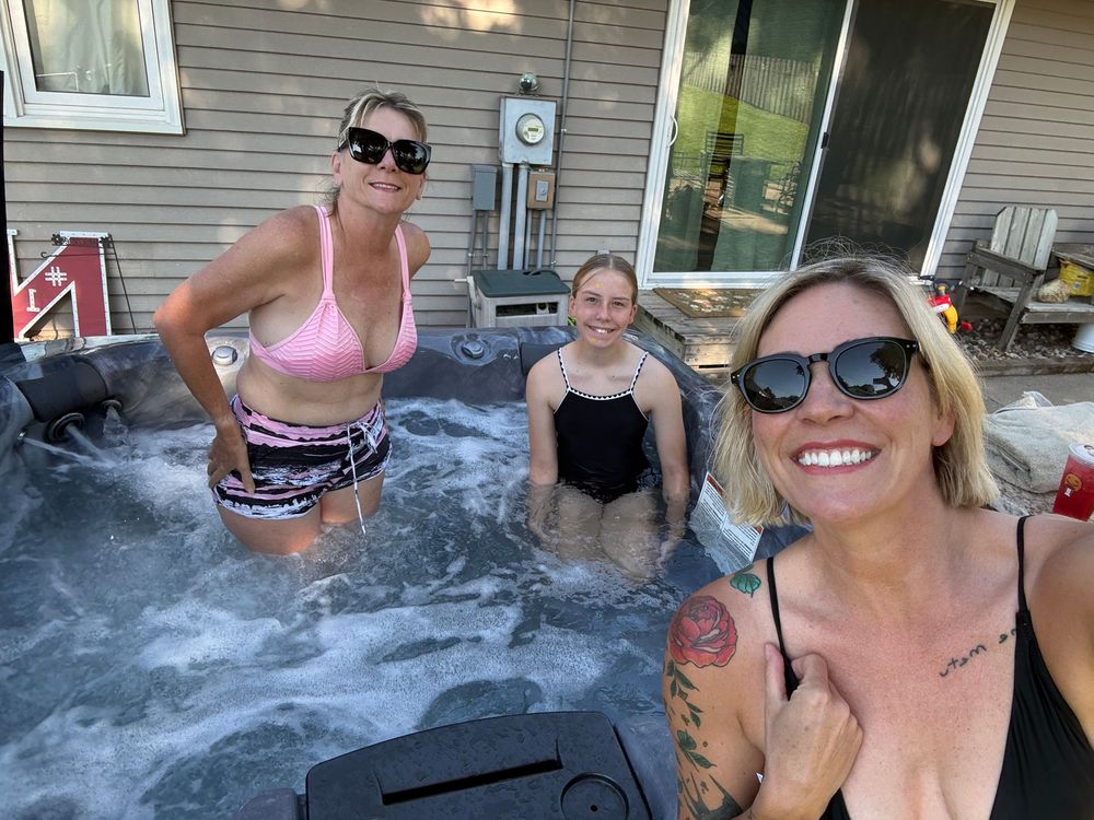 Three women enjoying a backyard hot tub on a sunny day, smiling and relaxing outdoors near a house.