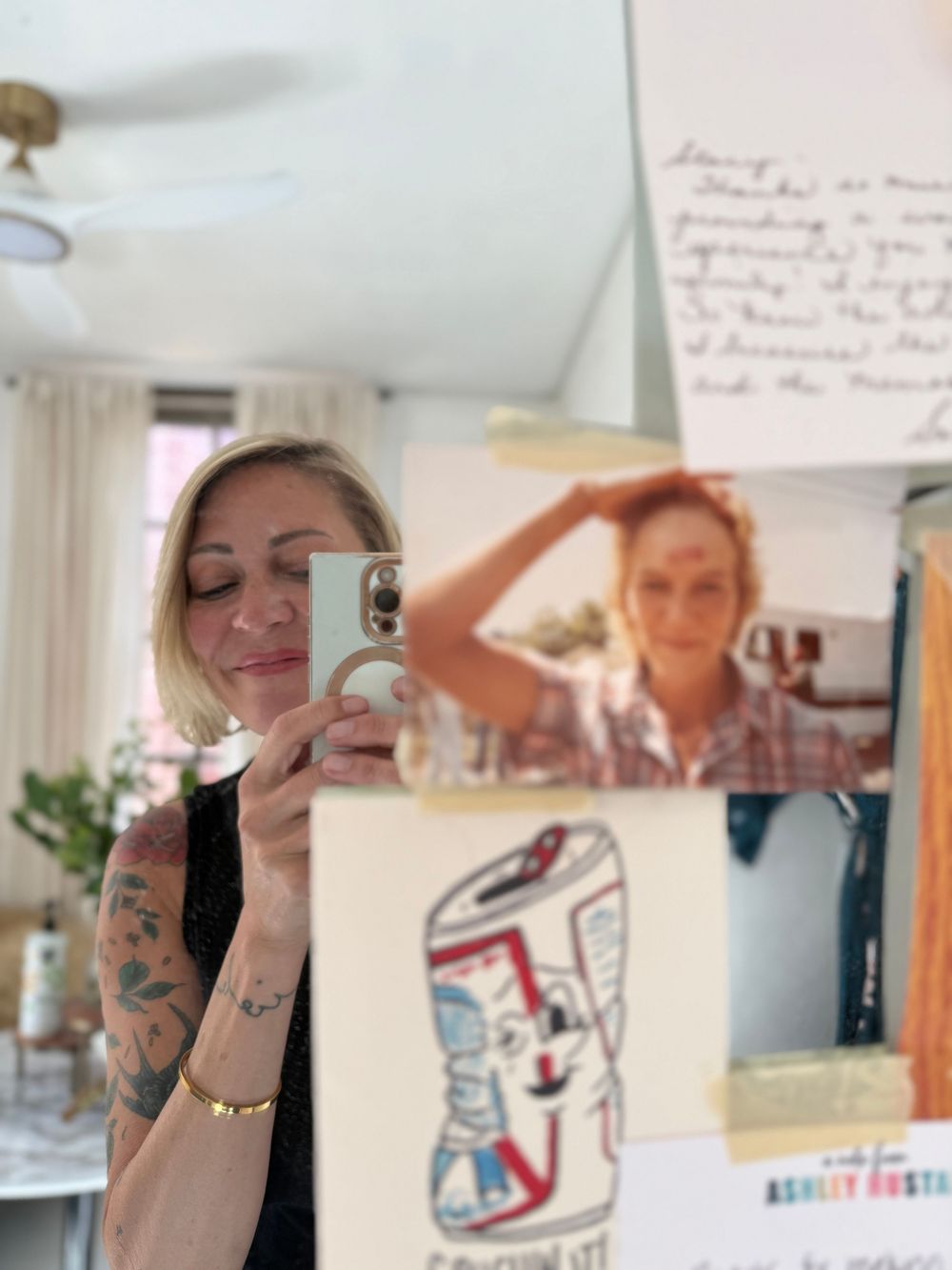 Tattooed blonde woman taking mirror selfie with phone, artwork and handwritten note visible in background.