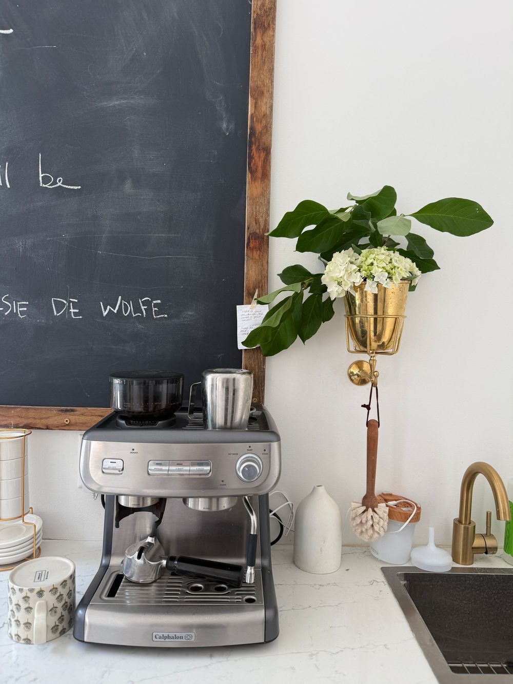 Stainless steel espresso machine on white counter beside chalkboard wall, gold wall vase with flowers, and brass faucet.