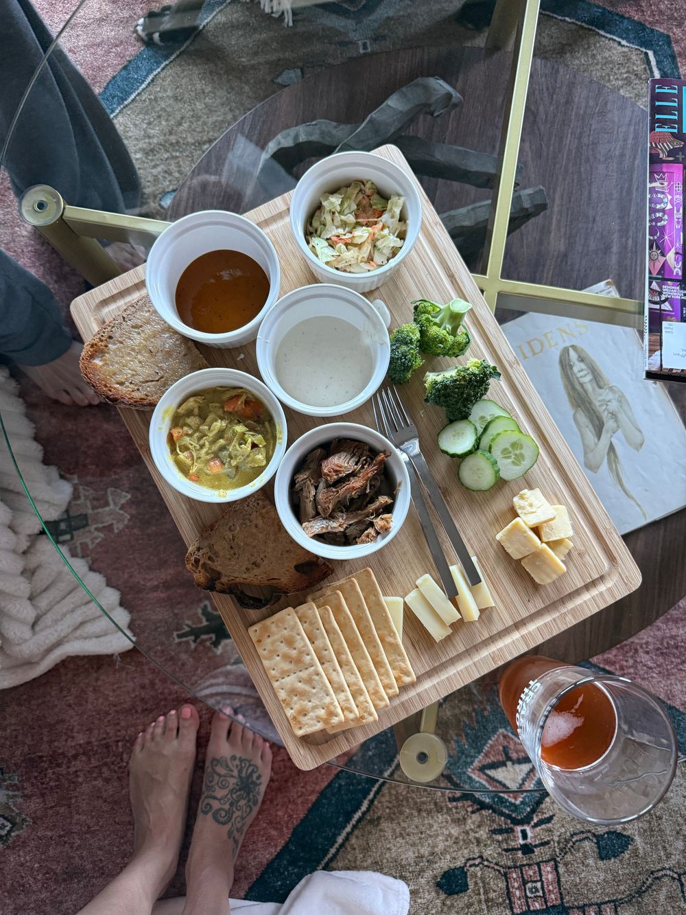 Overhead view of a wooden charcuterie board with cheese, crackers, soups, vegetables, and dips on a glass coffee table.
