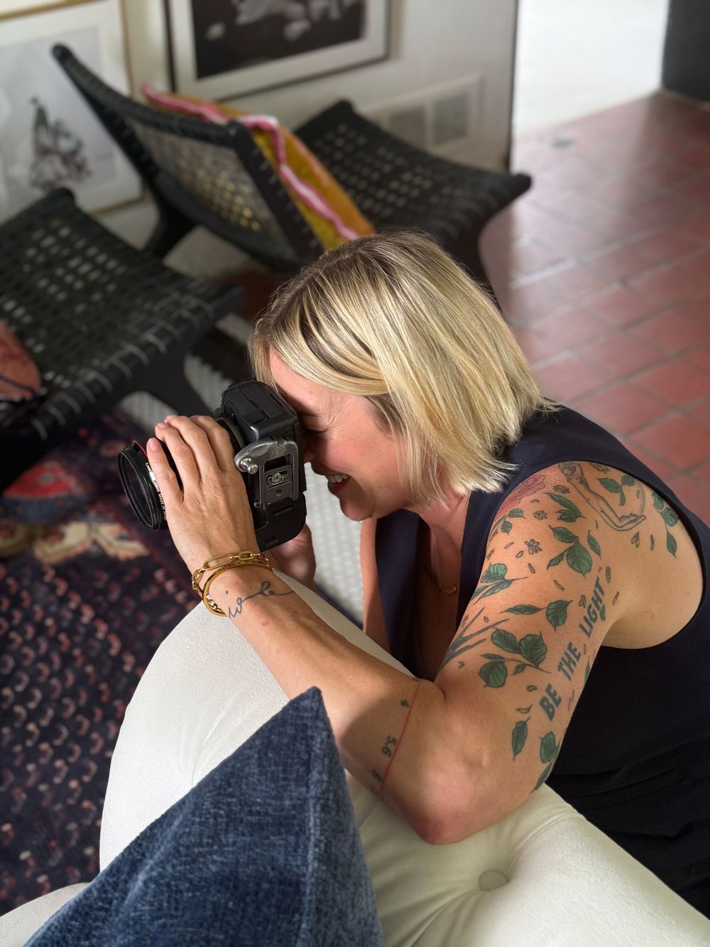 Blonde woman with floral arm tattoo photographing with a DSLR camera while sitting on a white couch indoors.