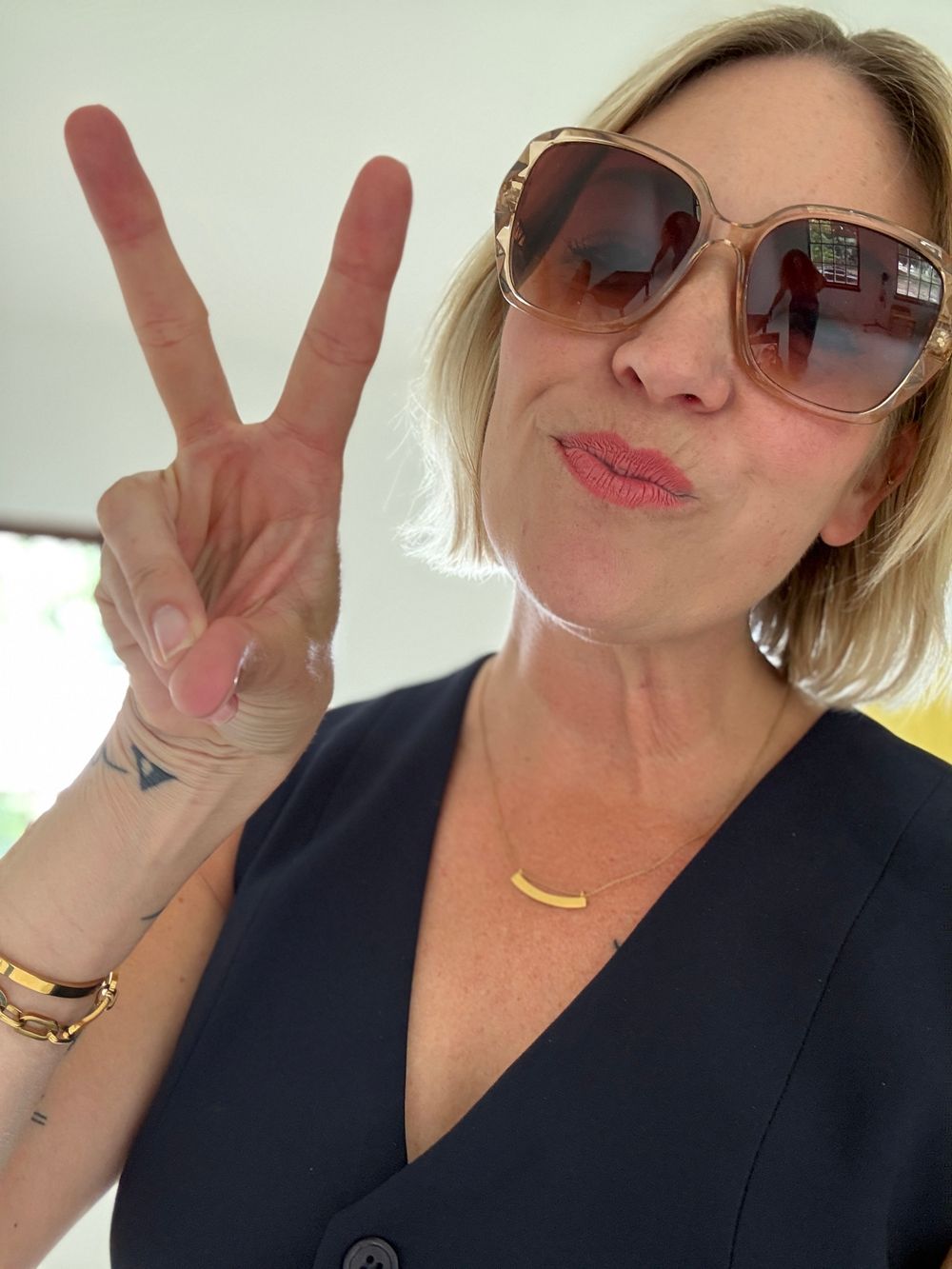 Woman in oversized sunglasses making a peace sign, wearing a navy top and gold necklace, posing for a selfie.