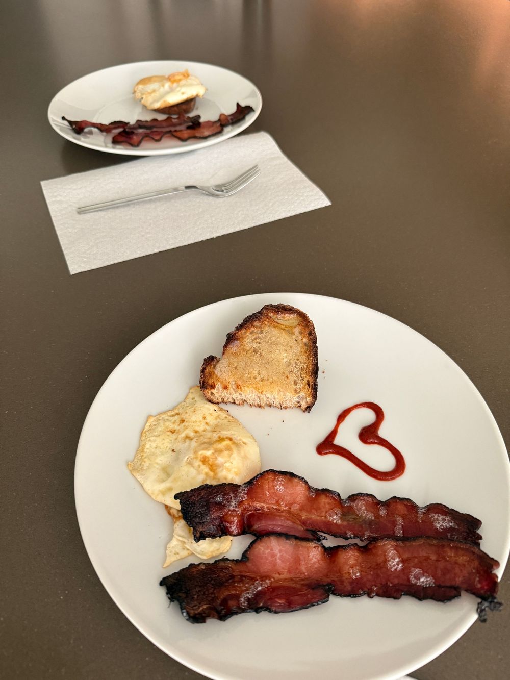 Two white plates with crispy bacon, fried eggs, toasted bread, and a ketchup heart on a gray table.