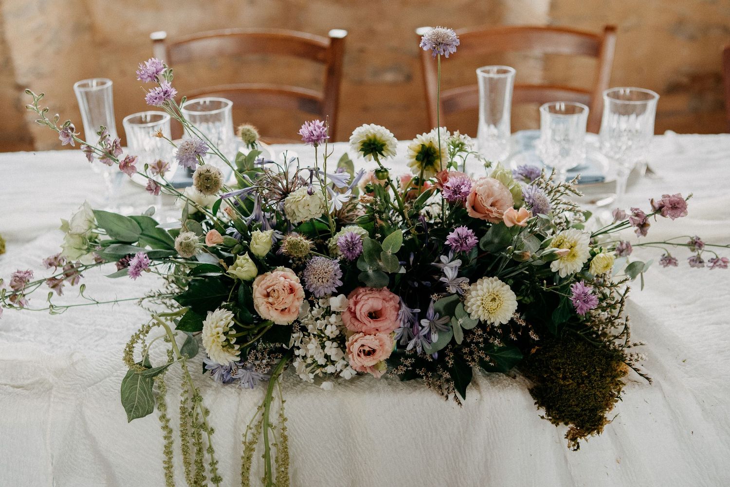 Lush wildflower wedding table centerpiece with pink roses, daisies, and greenery on a white linen tablecloth.