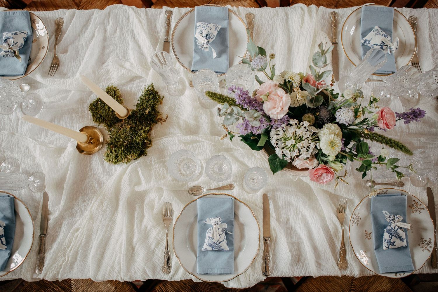 Elegant wedding reception table with dusty blue napkins, floral centerpiece of roses and hydrangeas, and white lace runner.