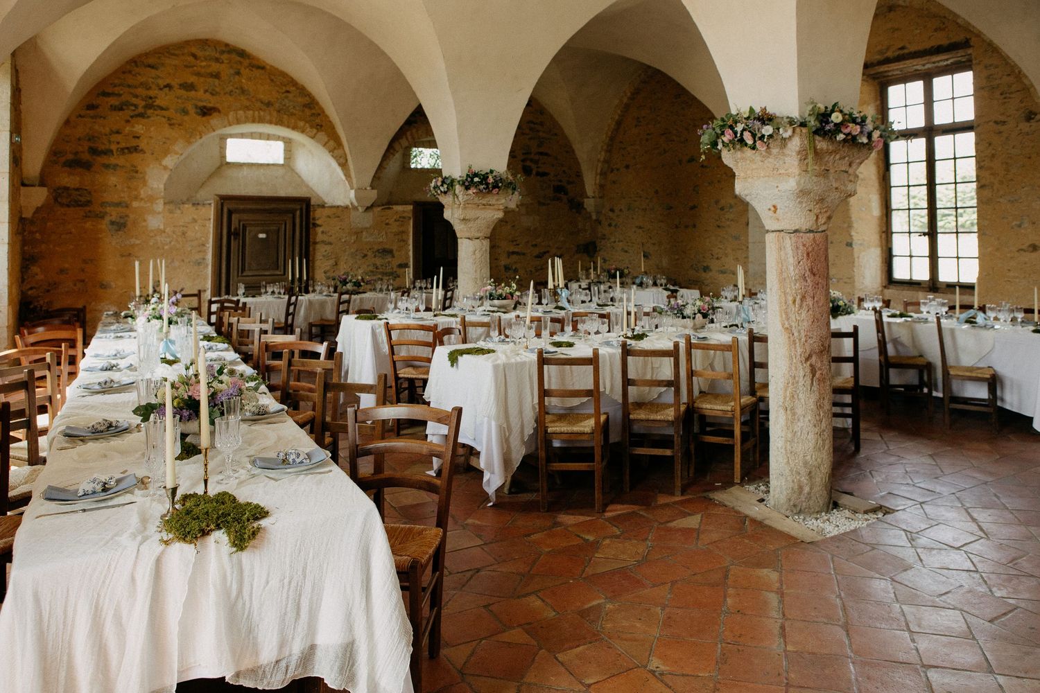Elegant wedding reception in a rustic stone vaulted hall with white linen tables, candles, and greenery centerpieces.