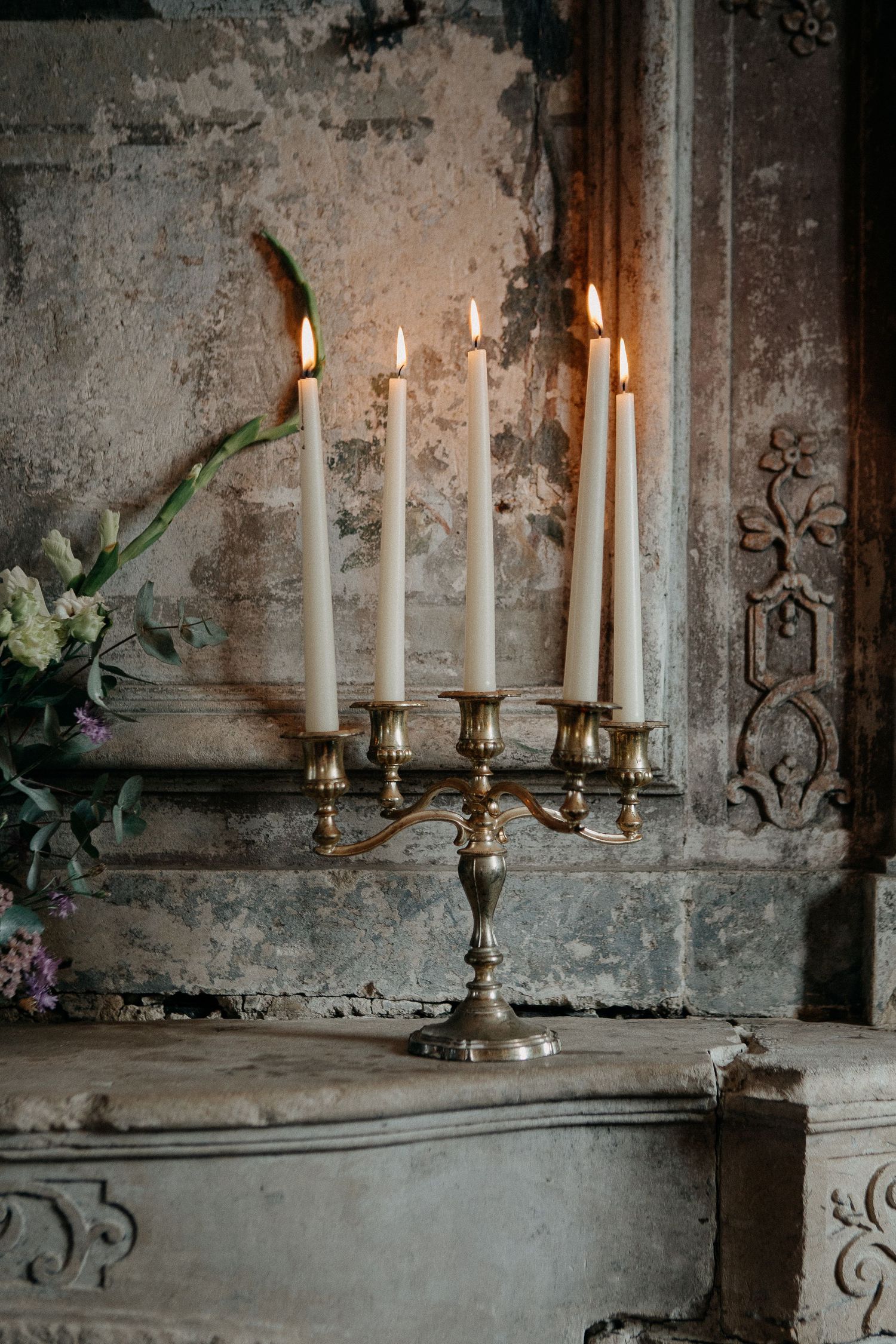 Ornate brass candelabra with five lit white taper candles on a vintage stone mantle in a rustic elegant setting.