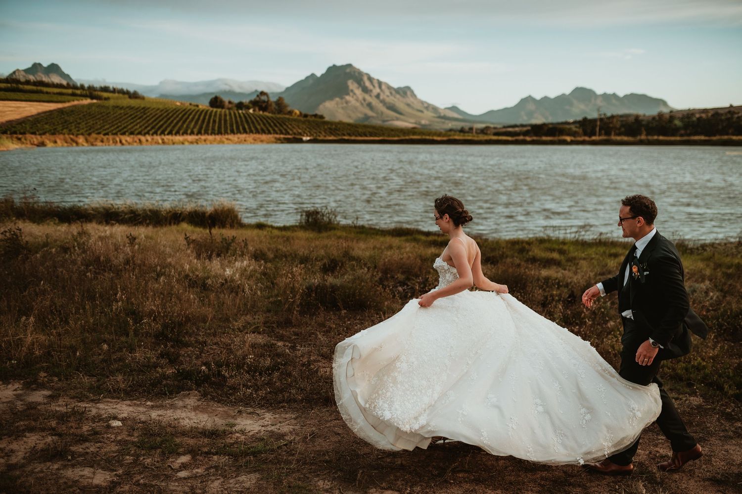 Bride and groom running near a lake with vineyard and mountain backdrop at a scenic outdoor wedding venue.