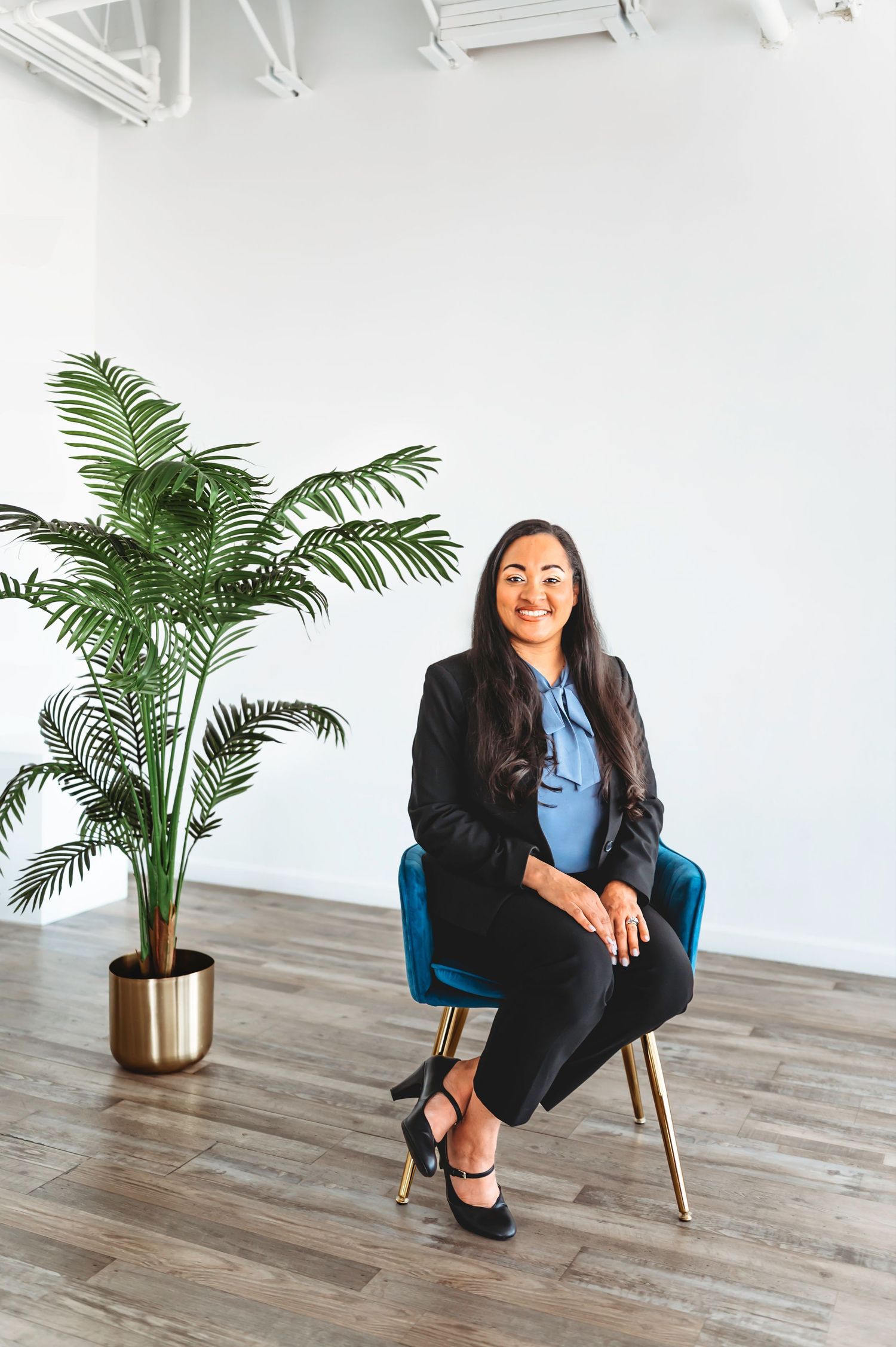 Professional woman in black blazer sitting in blue velvet chair beside tropical plant in bright studio.