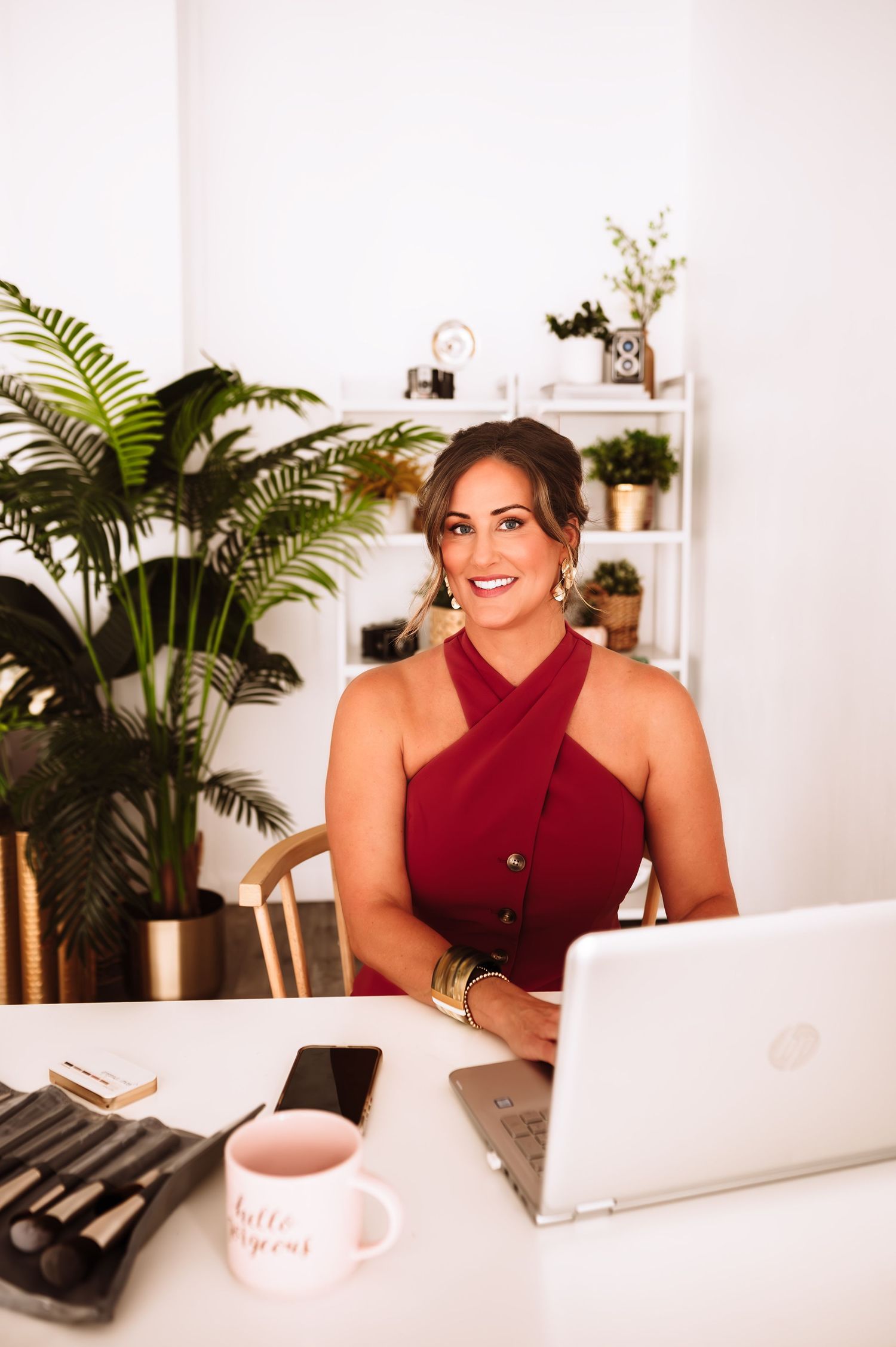 Woman in red dress smiling at desk with laptop, plants, and coffee mug in bright home office.