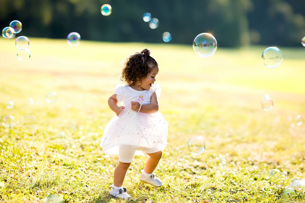 A toddler girl in a white dress chases soap bubbles in a sunny green field.