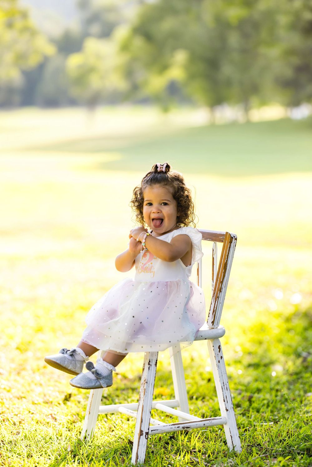 Toddler girl in white tutu dress sitting on vintage chair outdoors, clapping and laughing in sunny park.
