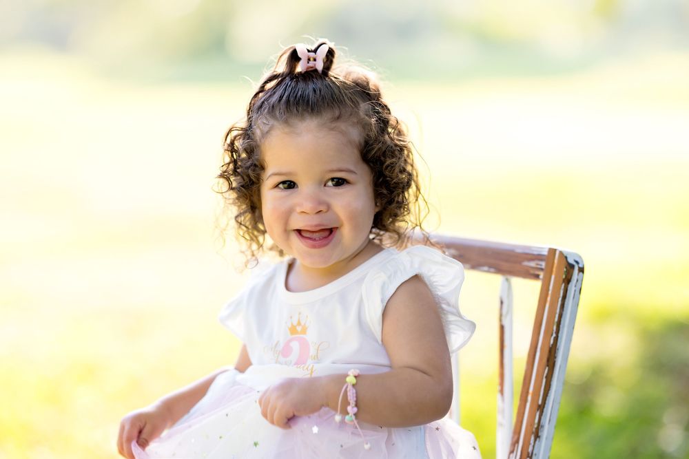Smiling toddler girl with curly hair and bow wearing white dress sits on chair in sunny outdoor setting.
