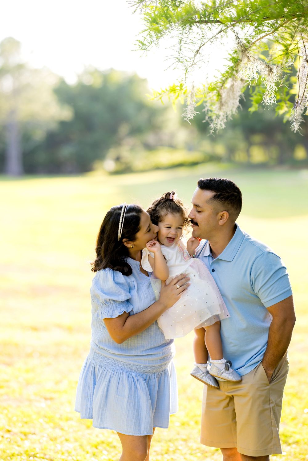 Happy family with parents in blue outfits holding their smiling toddler daughter in a sunny green park.