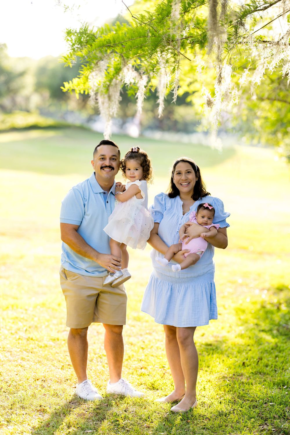 Happy family of four posing outdoors under flowering wisteria trees in a sunny green park setting.