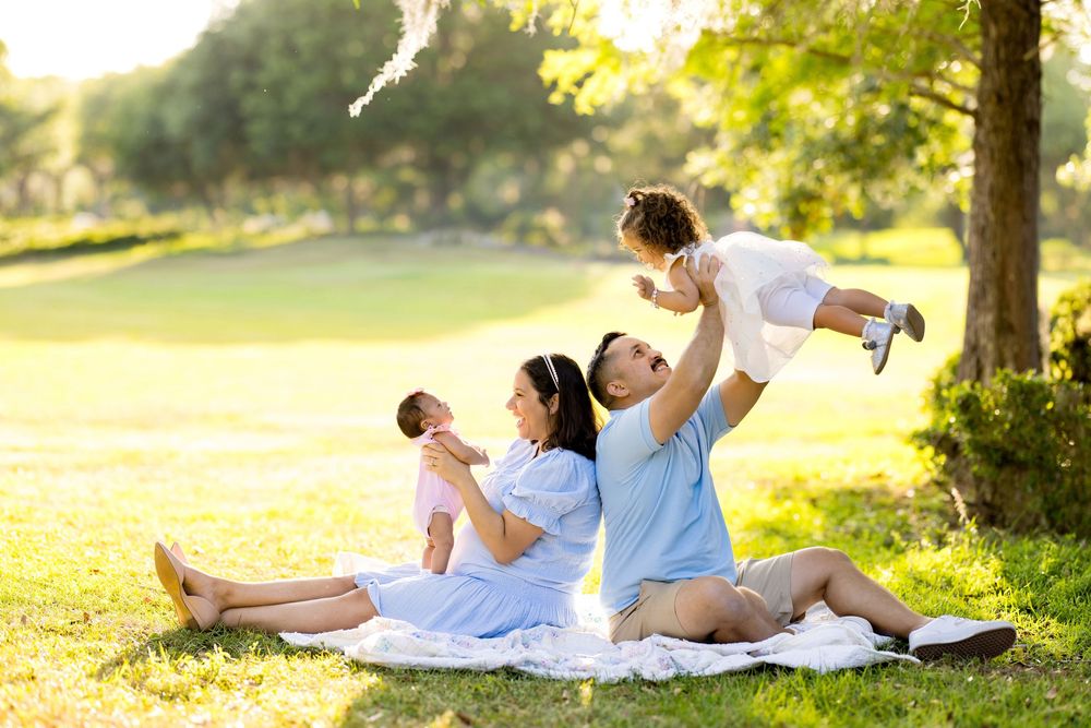 Happy family sitting on blanket in park, parents playing with two young children in sunny outdoor setting.