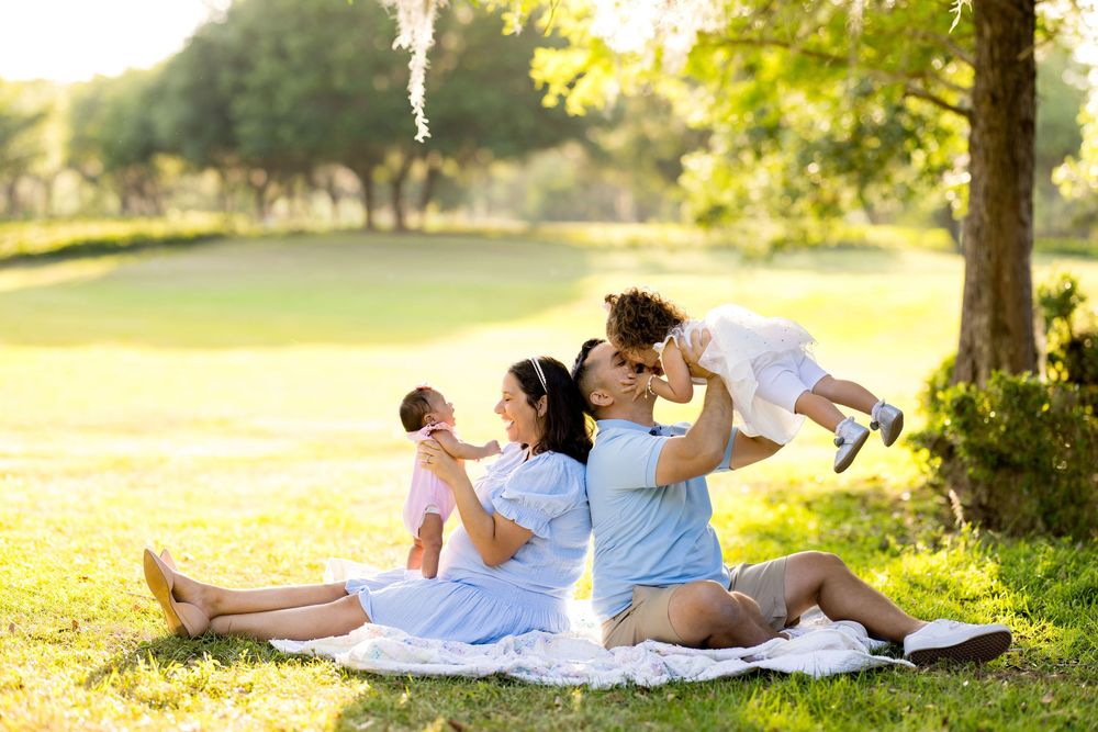 Family sitting on blanket in park, parents kissing while holding two young children in matching blue outfits on sunny day.
