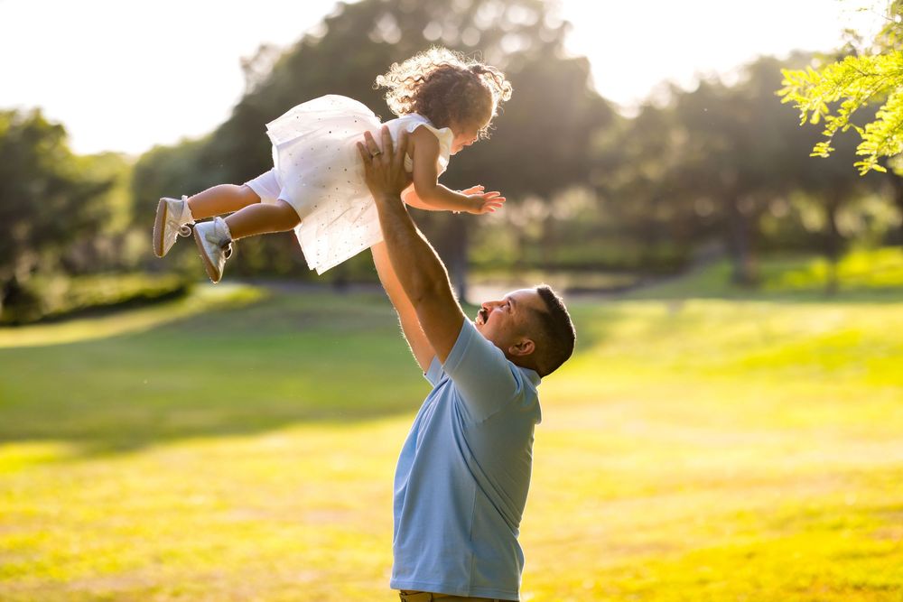 Father joyfully lifting his young daughter in a white dress high in the air in a sunny green park.