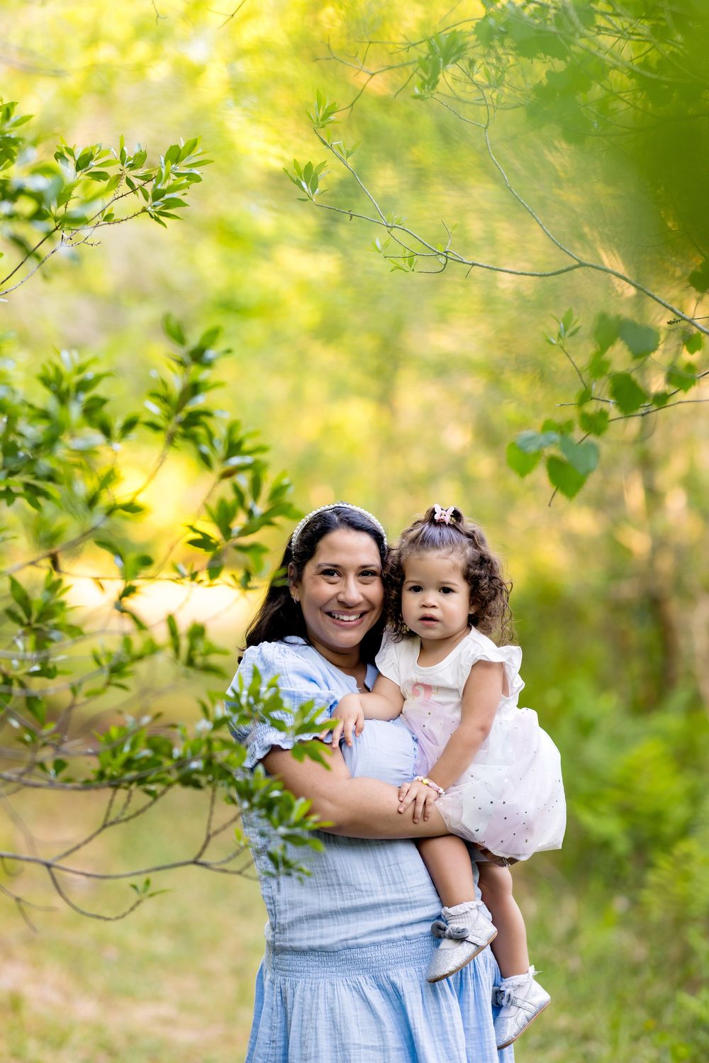 A smiling woman in a blue dress holds a toddler girl in a pink dress outdoors in a lush green park.