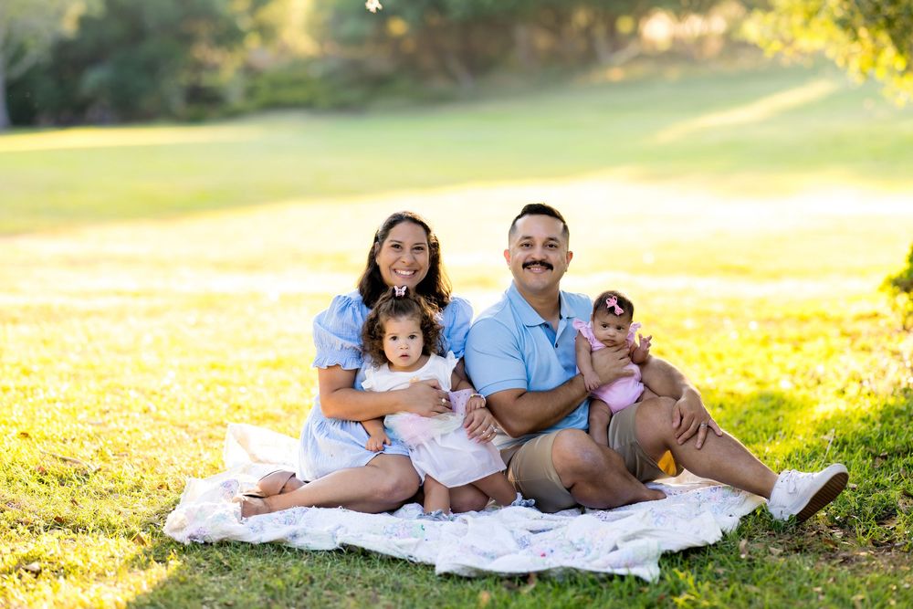 Family of four smiling on a blanket in a sunny park, parents holding two young children during an outdoor photo session.