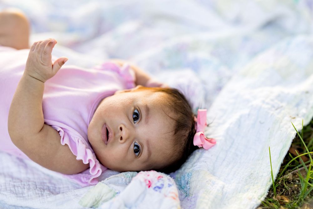 A cute baby girl in a pink onesie lying on a colorful blanket outdoors, looking up with bright curious eyes.