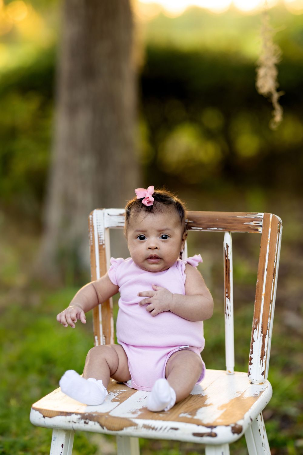 Adorable baby girl in pink onesie and bow sitting on a rustic white chair outdoors in a garden setting.