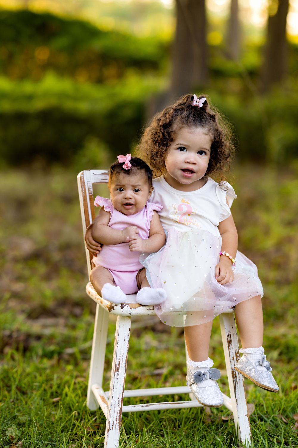 Two young sisters in pink outfits sit on a white chair outdoors, the older toddler holding her baby sister.