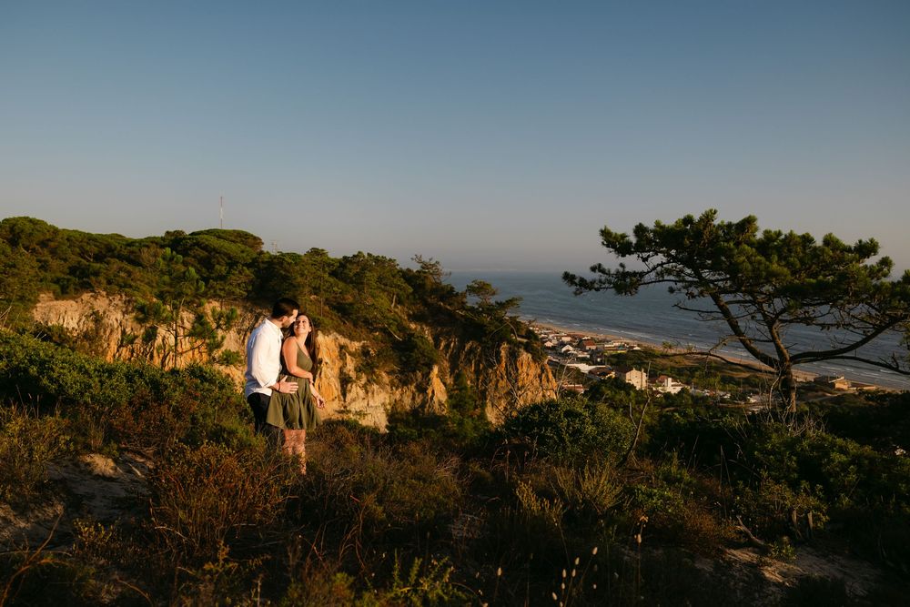 Couple embracing on a rocky hillside overlooking a coastal town at golden hour sunset.