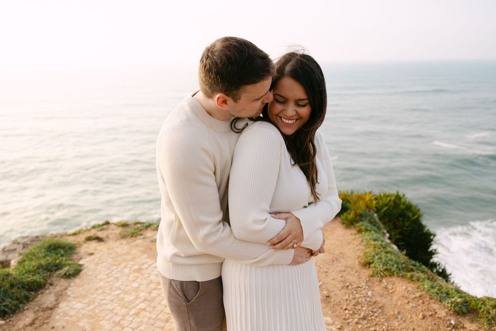 Couple embracing on coastal cliff, woman in white dress smiling, man in beige sweater, ocean backdrop at golden hour. Azenhas do Mar
