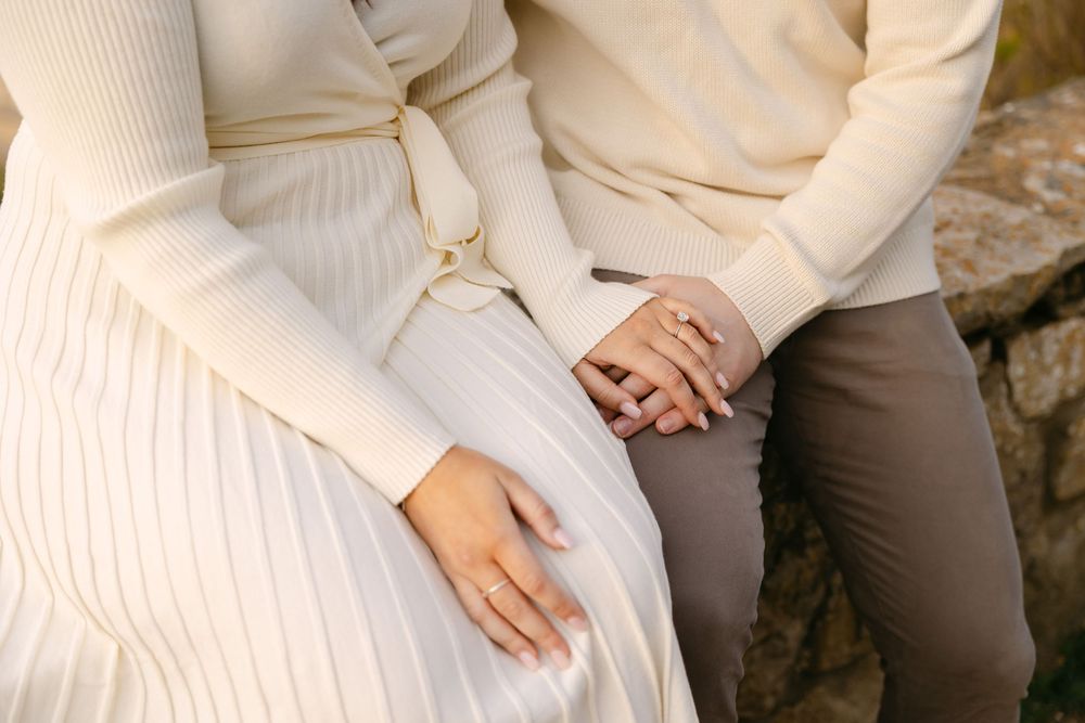 Couple holding hands while sitting together, wearing cream and beige outfits, with an engagement ring visible. Azenhas do Mar
