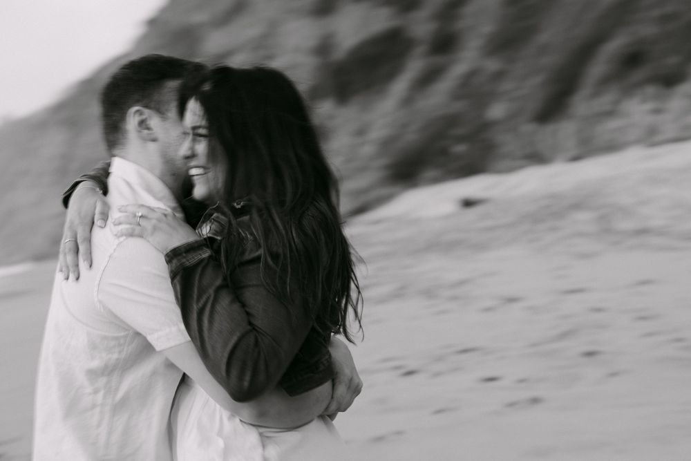 Black and white photo of a couple embracing and smiling on a sandy beach with rocky cliffs in the background. Praia da Adraga