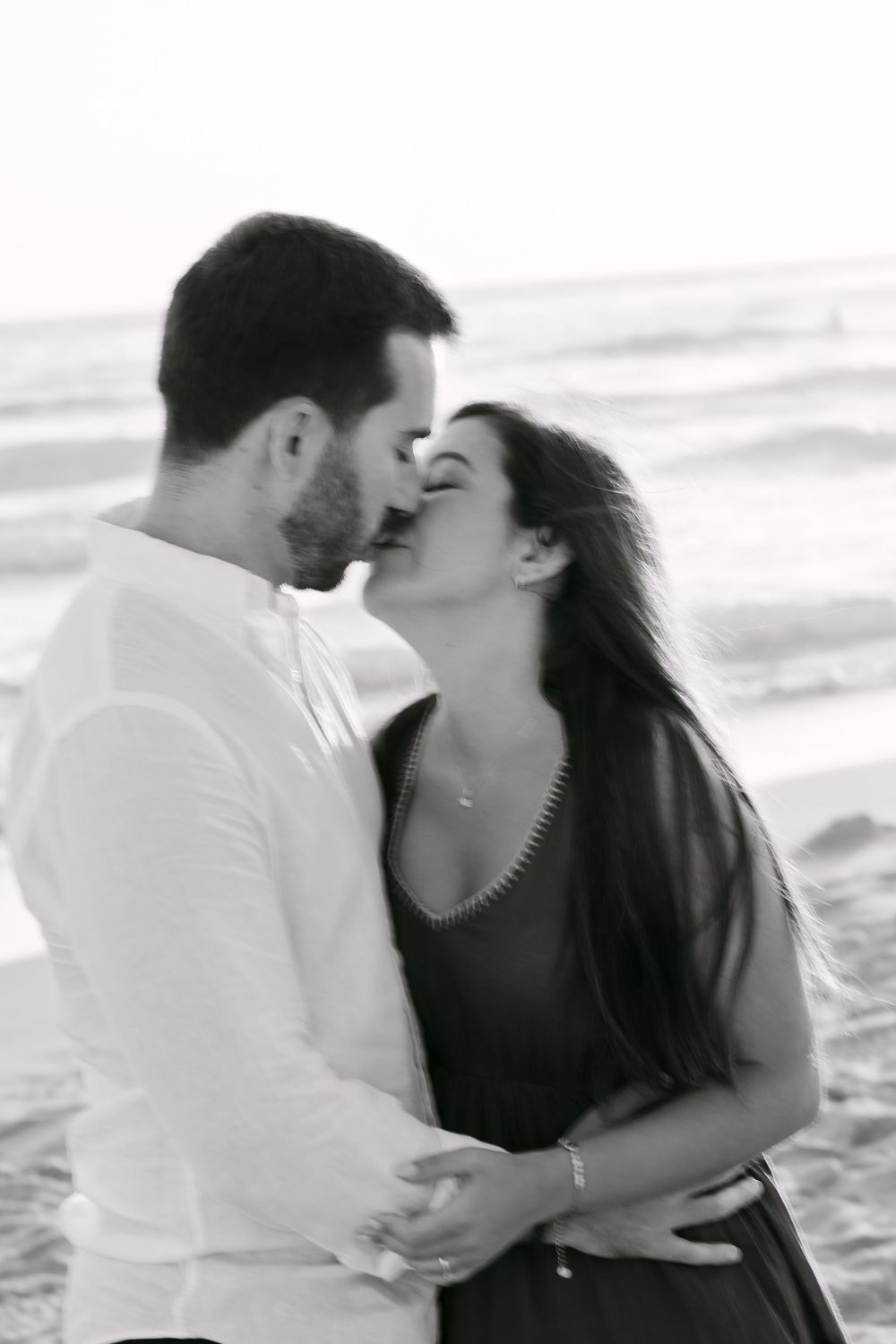 Black and white photo of a romantic couple sharing a kiss on the beach with ocean waves in the background. Praia da fonte da telha