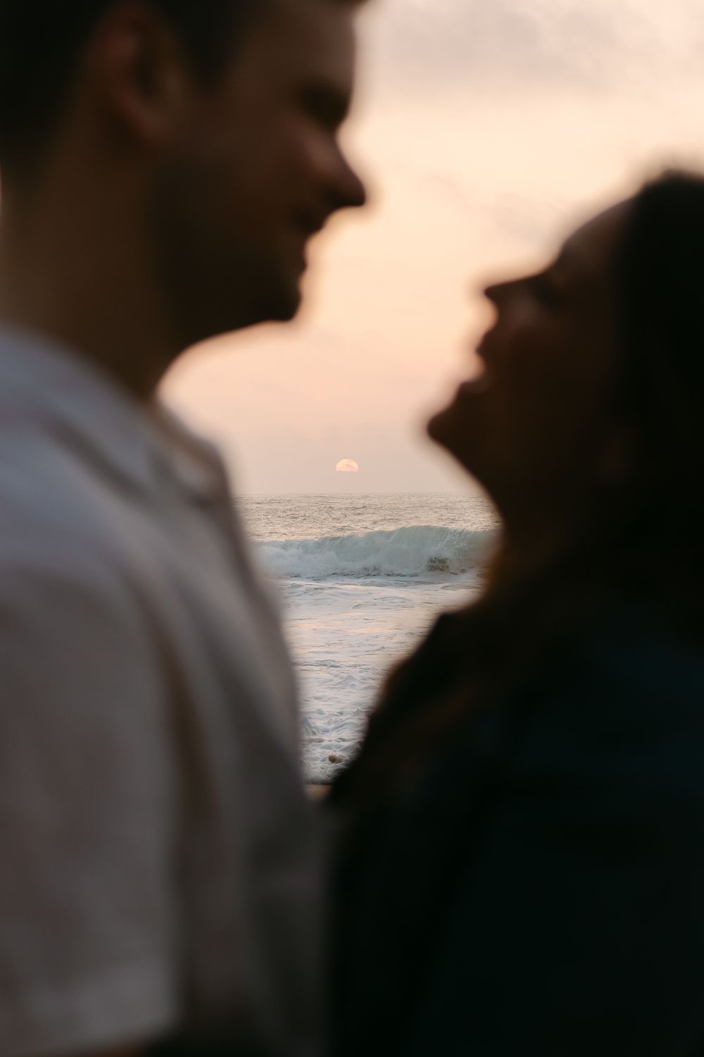 Couple sharing a romantic moment at sunset by the ocean with waves and glowing horizon in the background. Praia da Adraga