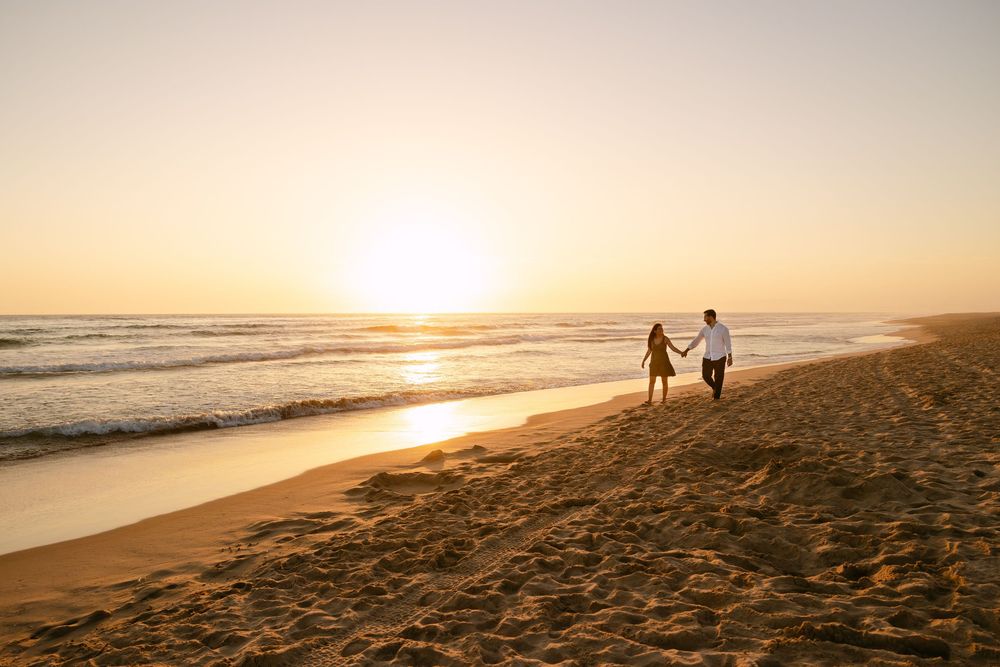 Couple holding hands walking on a sandy beach at golden sunset with ocean waves in the background. Praia da fonte da telha