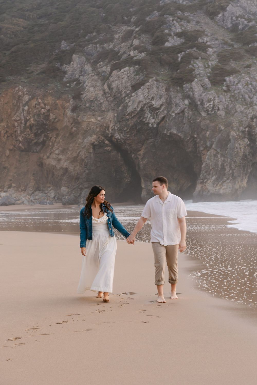 Couple holding hands walking barefoot on sandy beach with rocky cliffs in background, woman in white dress and teal jacket. Praia da Adraga
