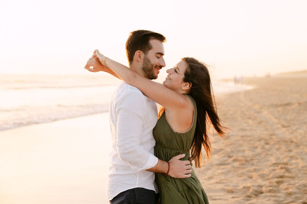 Couple embracing romantically on a sandy beach at golden sunset, woman in green dress with hair blowing in the wind. Praia da fonte da telha