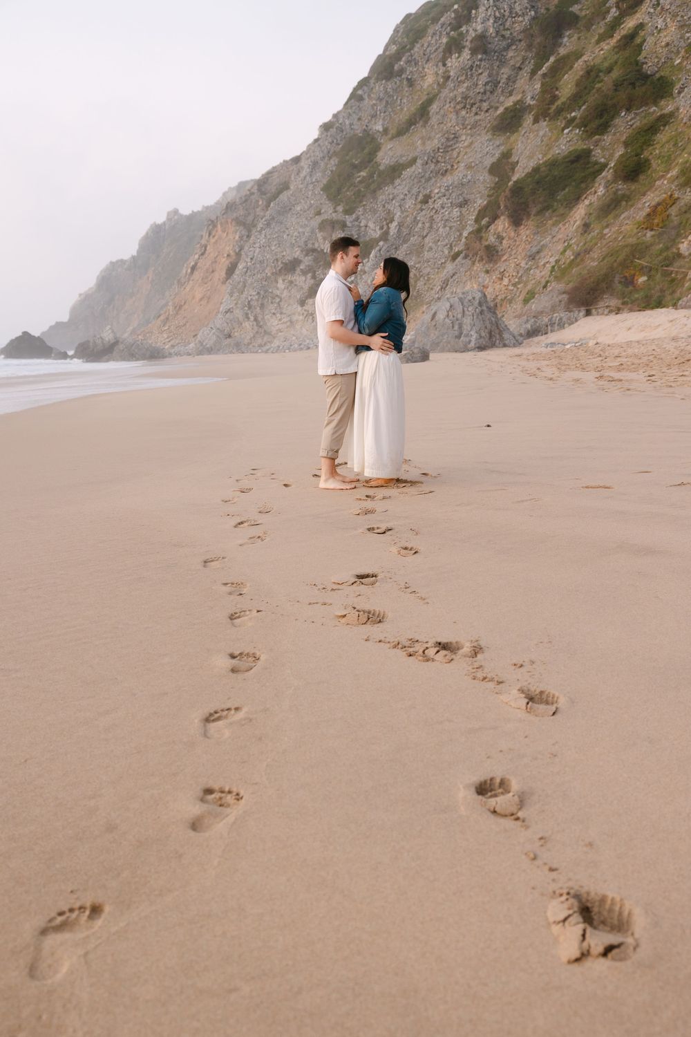 Couple embracing on sandy beach with footprints trail, rocky cliffs in background on overcast day. Praia da Adraga