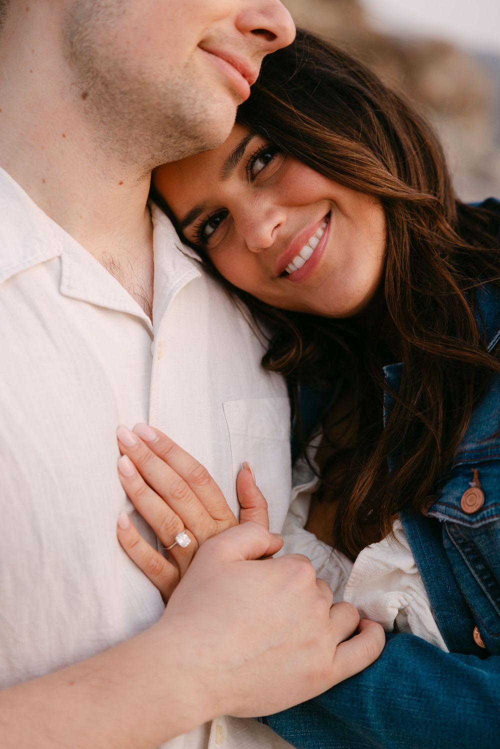Smiling woman in denim jacket hugs man, showing off engagement ring, in a romantic close-up portrait. Praia da Adraga