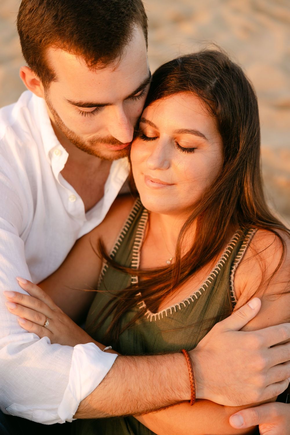 A couple embraces tenderly on a beach, man in white shirt kissing woman's temple at golden hour.
