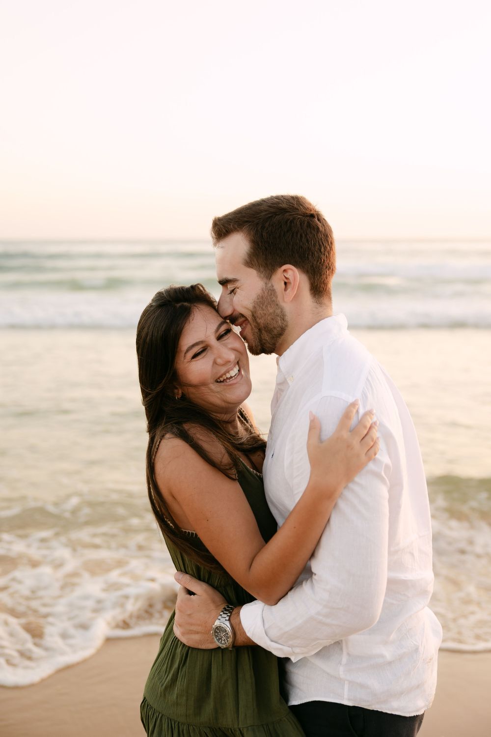 A couple embraces and laughs together on a sandy beach at sunset, with ocean waves in the background. Praia da fonte da telha