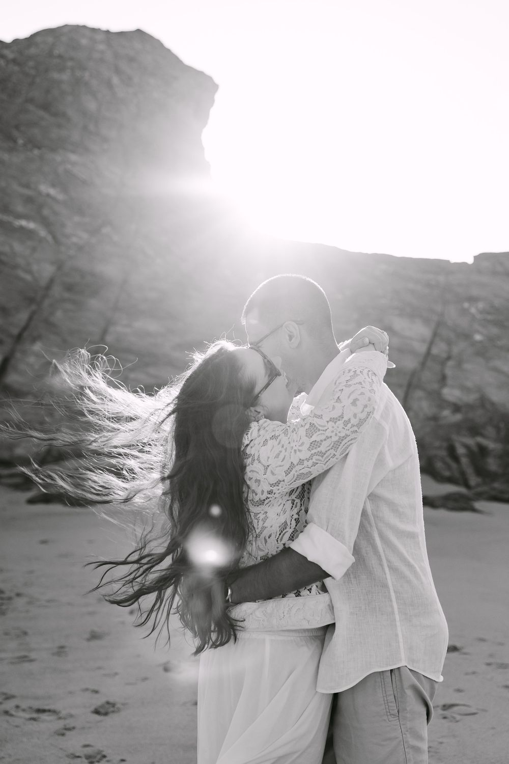 Black and white photo of a couple kissing on a beach with wind-blown hair and sun flare behind rocky cliffs.Praia da samouqueira, Porto Covo
