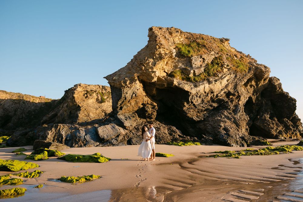 Couple embraces on sandy beach with dramatic rocky cliffs and green moss in golden hour sunlight. Praia da samouqueira, Porto Covo