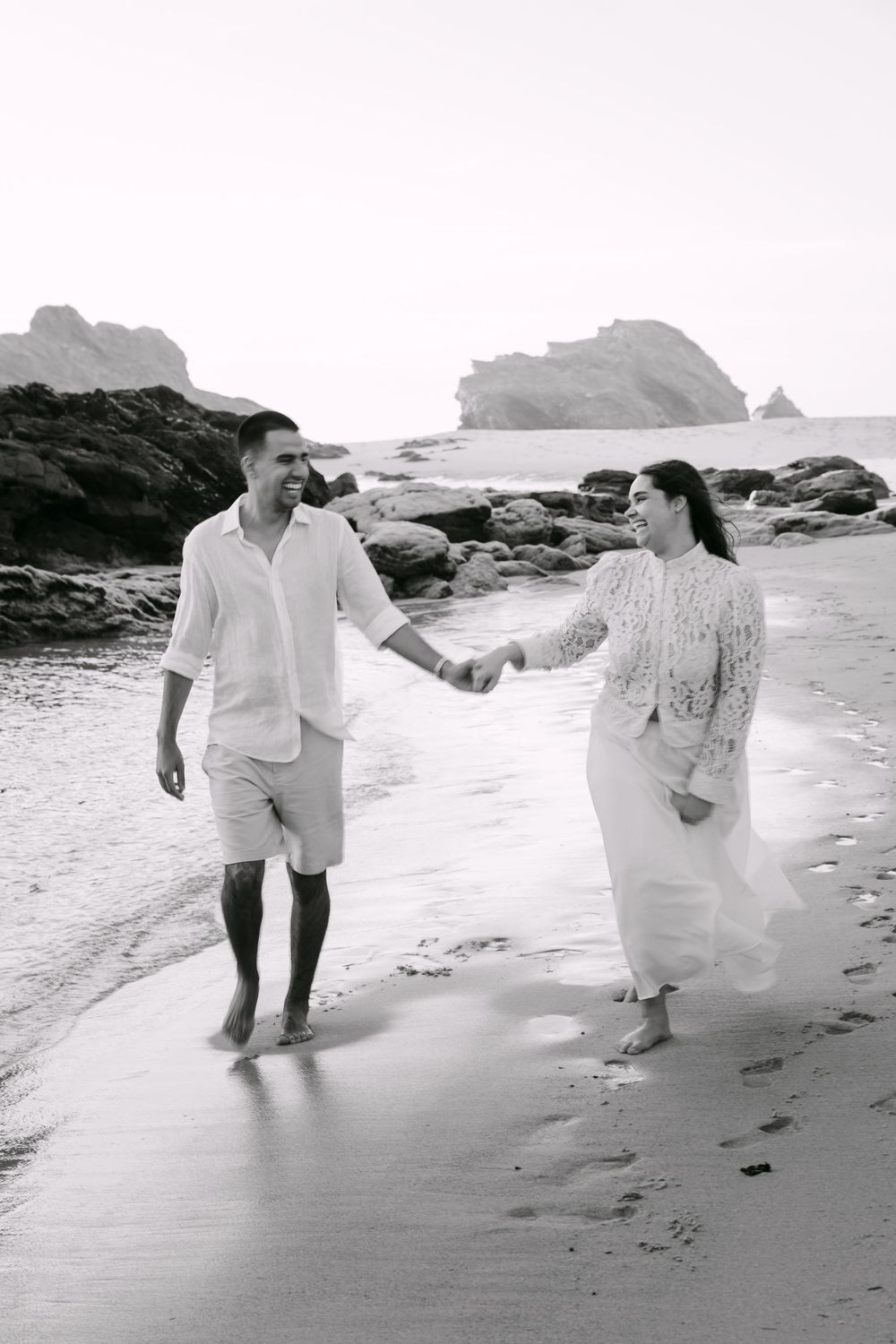 Black and white photo of a couple holding hands and walking barefoot on a rocky beach in white outfits. Praia da samouqueira, Porto Covo