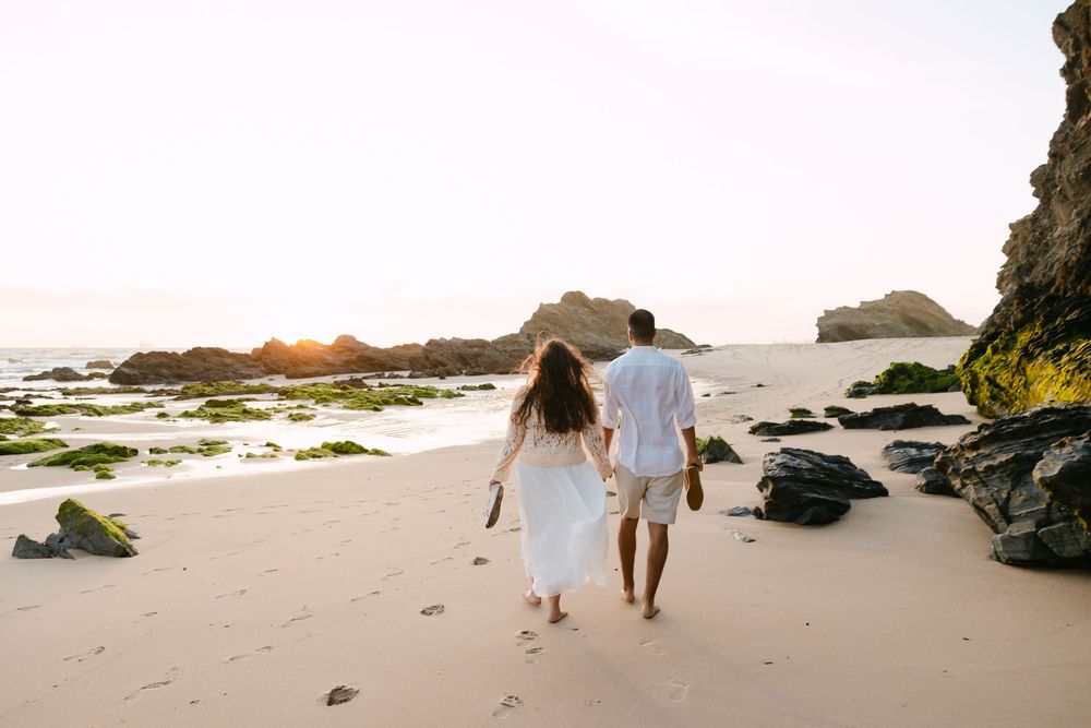Couple in white outfits walking hand in hand on a sandy beach at sunset with rocky cliffs in the background. Praia da samouqueira Porto Covo