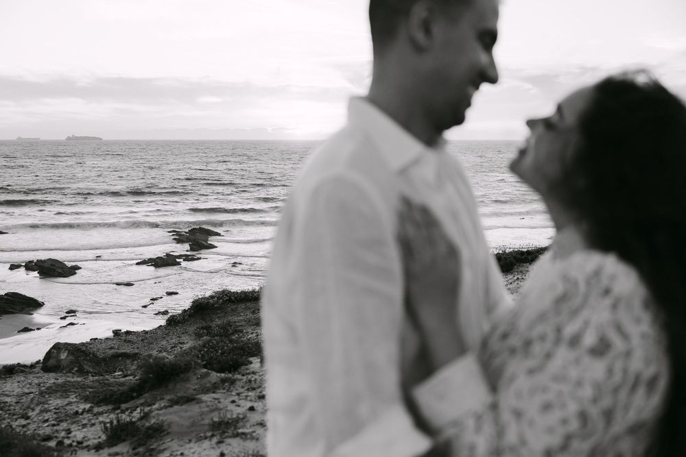 Black and white photo of a couple embracing on a rocky beach at sunset, gazing lovingly at each other. Praia da samouqueira, Porto Covo